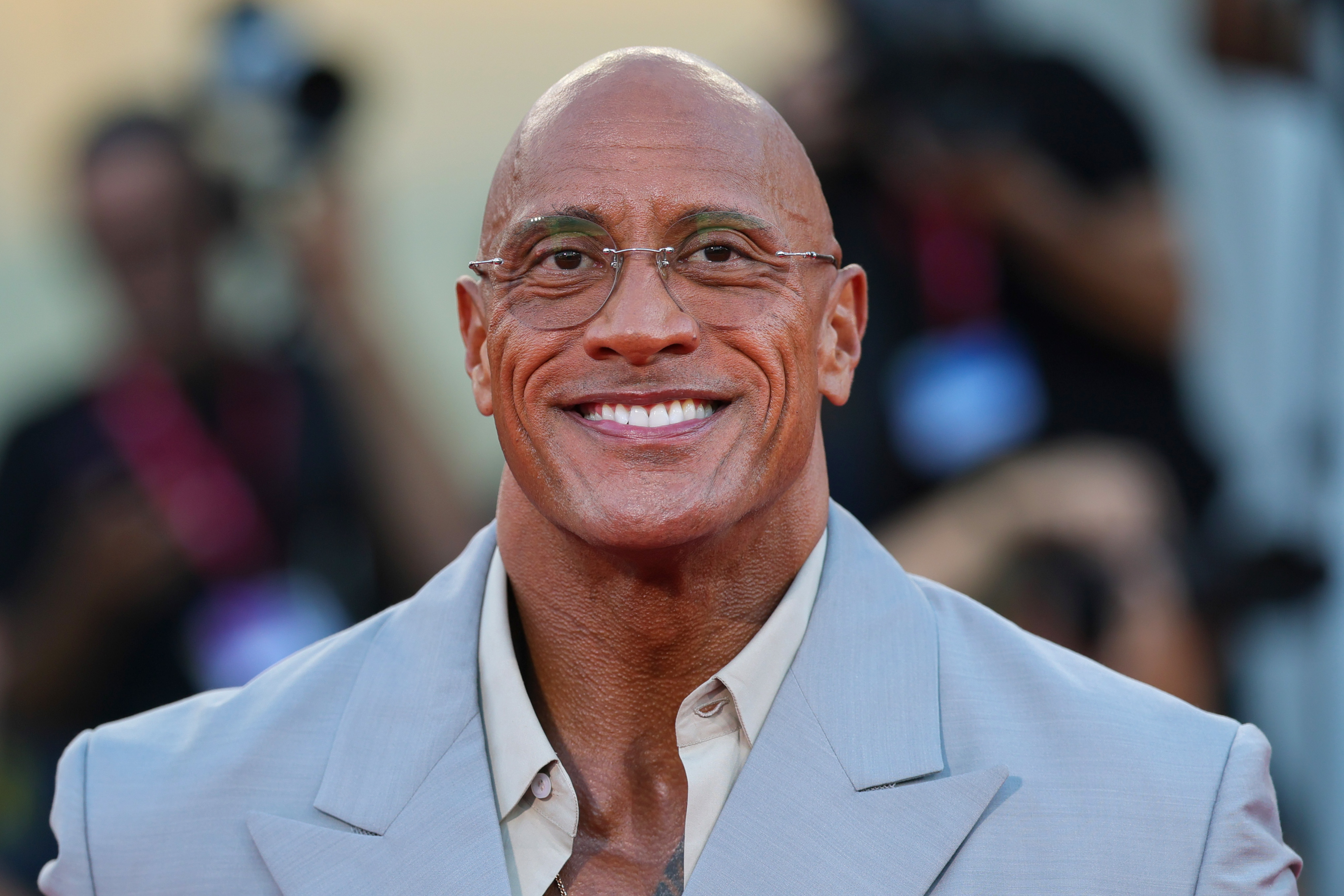 Dwayne Johnson sonriente, viste un traje claro en un evento.