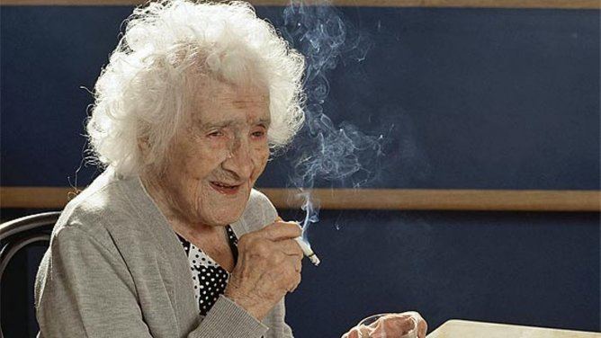 Mujer mayor sonriente con cabello rizado, sosteniendo un cigarrillo.