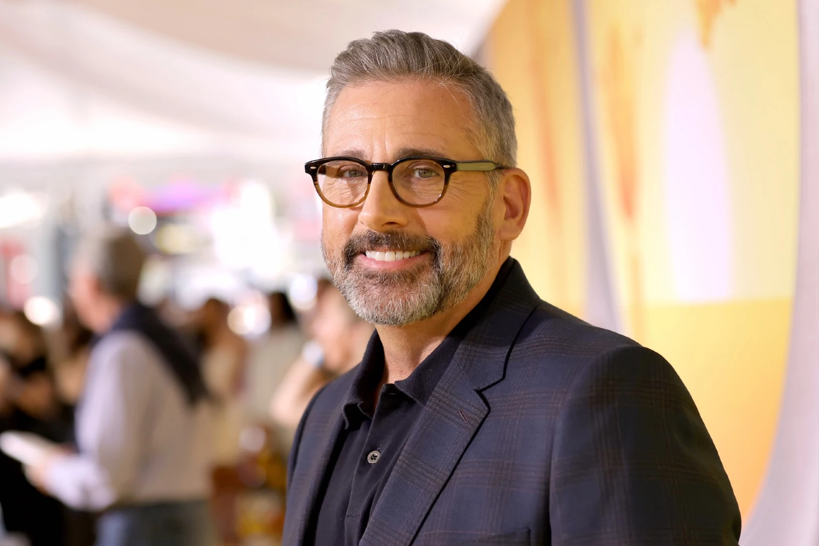 Steve Carell sonriendo en un evento, con fondo borroso.